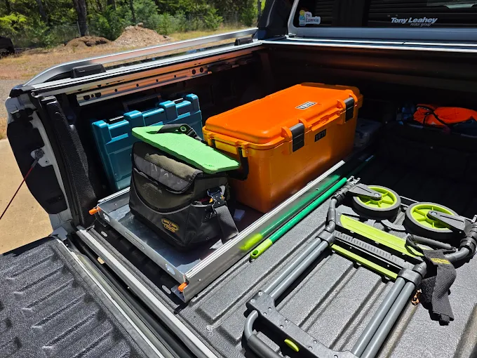 Pickup truck bed with orange case, green-handled tools, bags, blue case, and folding dolly.