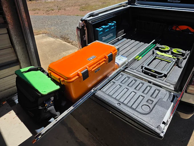 Pickup truck bed with organized tools, cooler, bags, poles, and foldable equipment on a sliding tray.